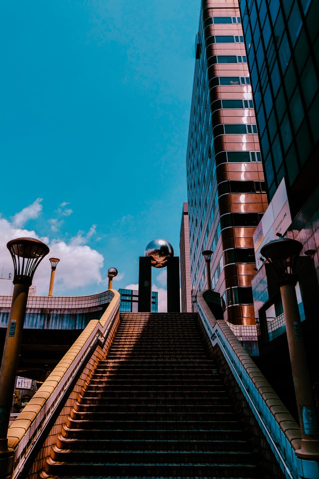A long flight of steps climbs between street lamps towards a glass tower under a hard blue sky.