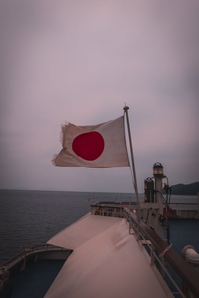 A flag flies from the bow of a ferry over open grey water, a low headland on the horizon.