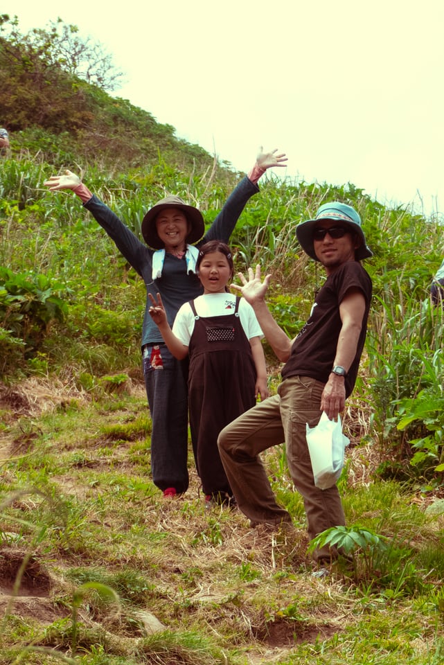 Three people stand on a green hillside above the sea with their arms raised, two of them in wide sun hats.