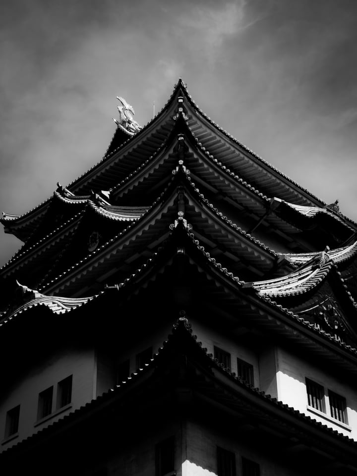 In black and white, the tiered roofs of a castle keep rise against heavy cloud.